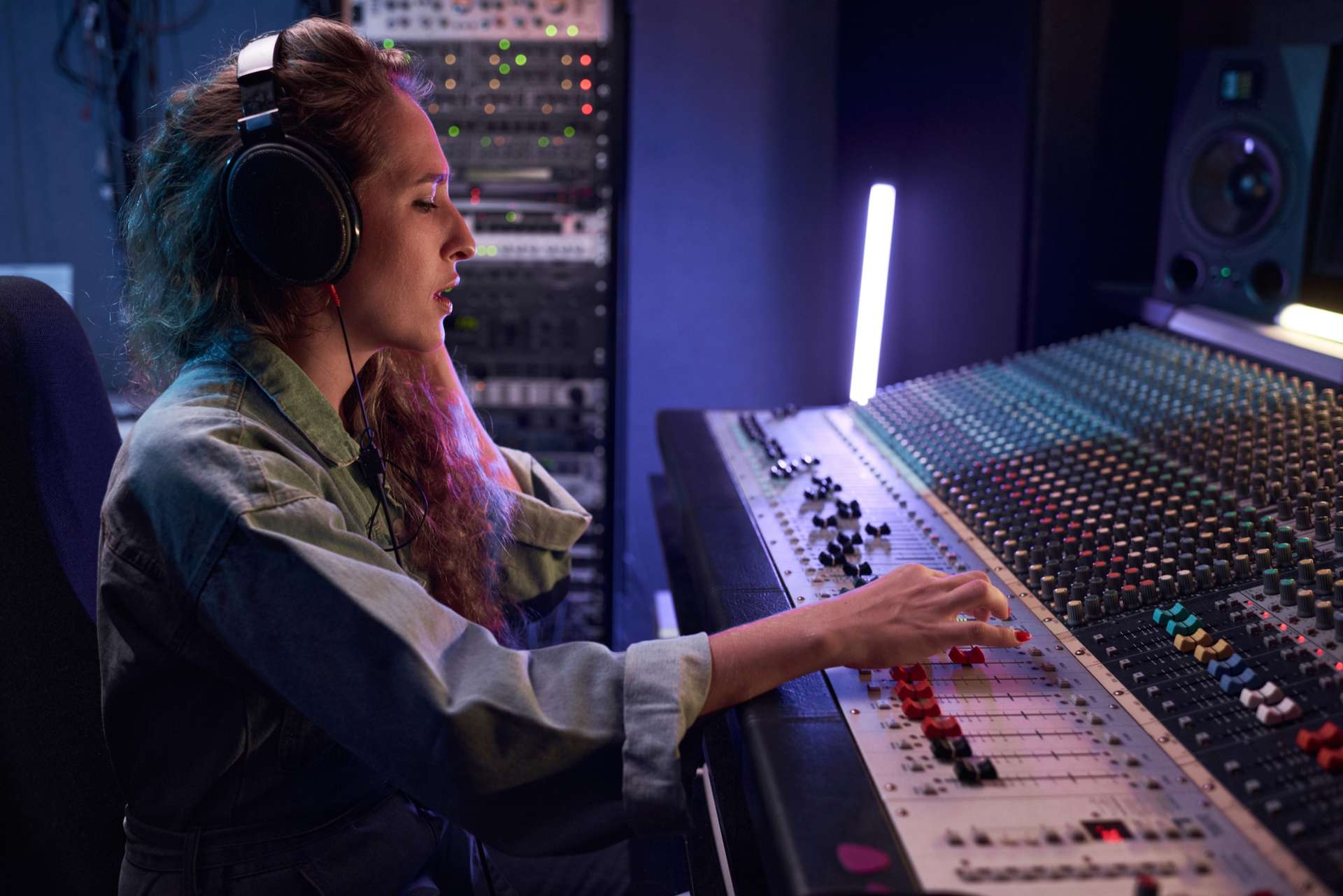 Mixing at an analog console: headphones on, a hand on the faders, racks of outboard gear behind.
